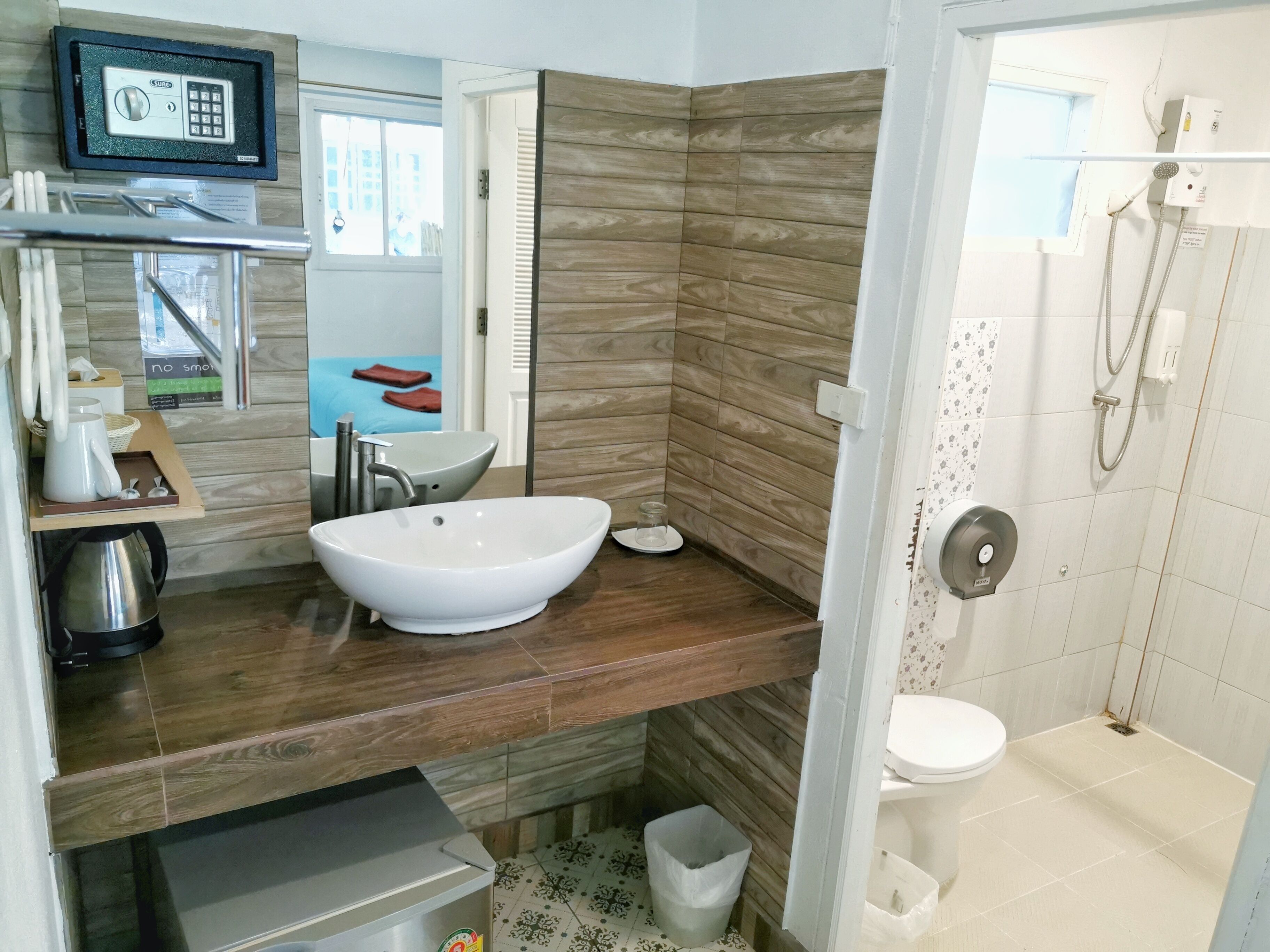 Een moderne badkamer met een groot wit wastafel, douche en toilet. Er is een houten opbergmeubel met spiegel en een klein plankje met toiletartikelen.