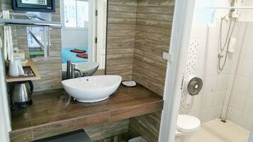 Een moderne badkamer met een groot wit wastafel, douche en toilet. Er is een houten opbergmeubel met spiegel en een klein plankje met toiletartikelen.