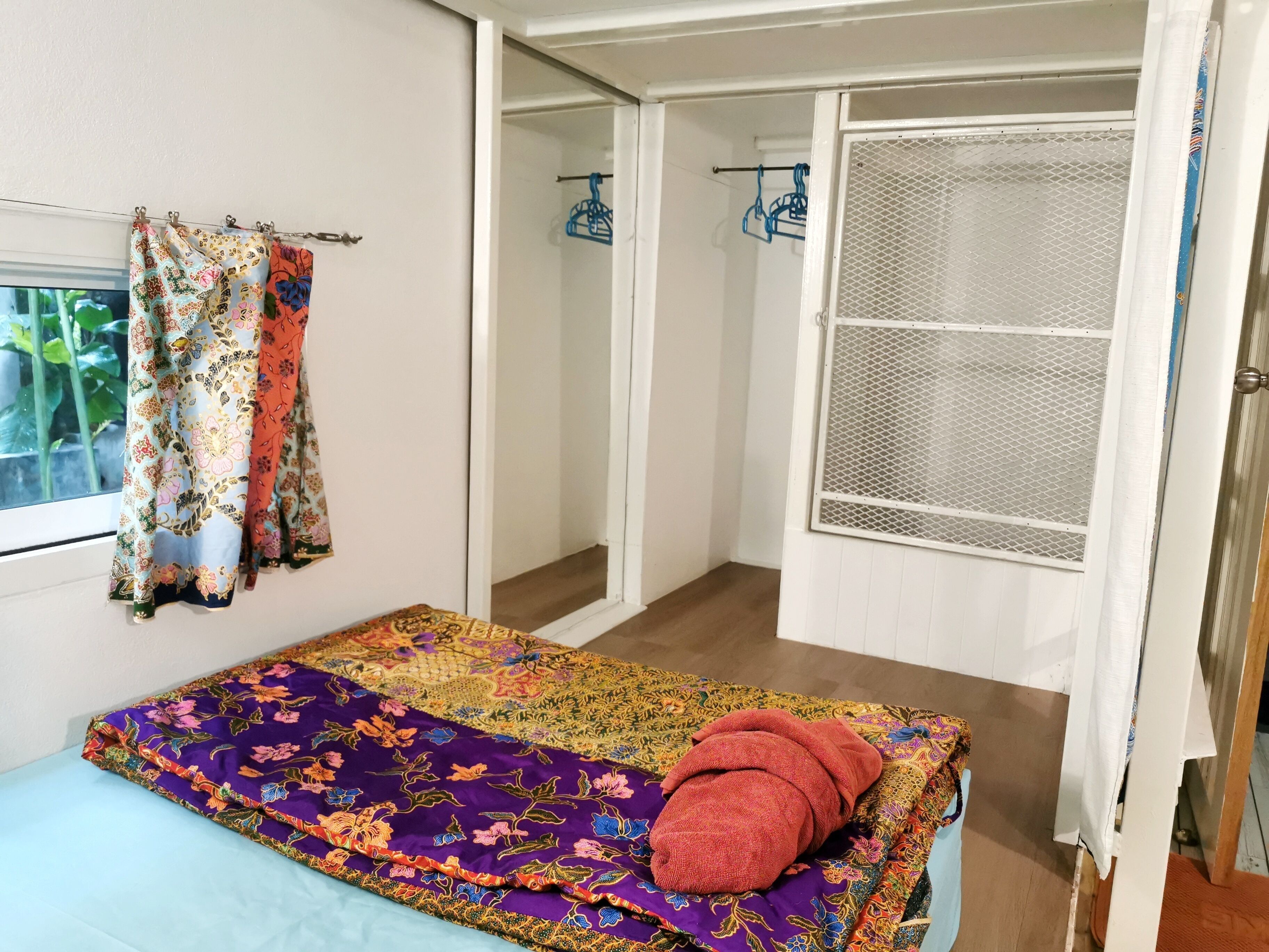 A room with a bed, a window with curtains, a door with a mesh screen, and a hallway with a clothes rack.
