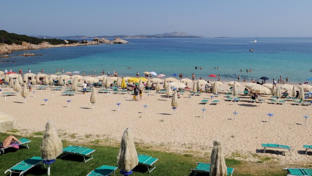 Una spiaggia con lettini e ombrelloni, un mare cristallino e montagne in lontananza.