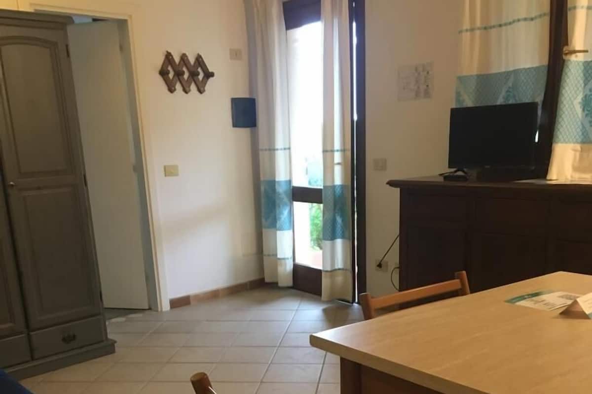 Un coin repas avec une table et des chaises en bois, un téléviseur posé sur un meuble, et une vue sur l’extérieur par une fenêtre ornée de rideaux.