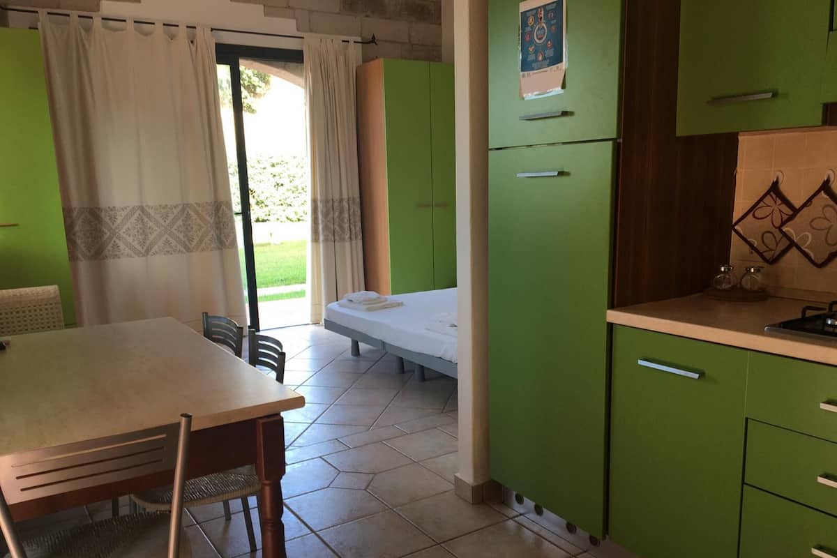 Une pièce comprenant un lit, une table à manger avec des chaises, un coin cuisine avec des meubles verts et une vue sur l’extérieur grâce à une porte vitrée.