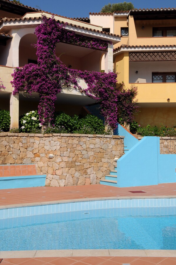Zona de piscina con un muro de piedra y una escalera azul que conduce a un edificio con un balcón cubierto de flores moradas.