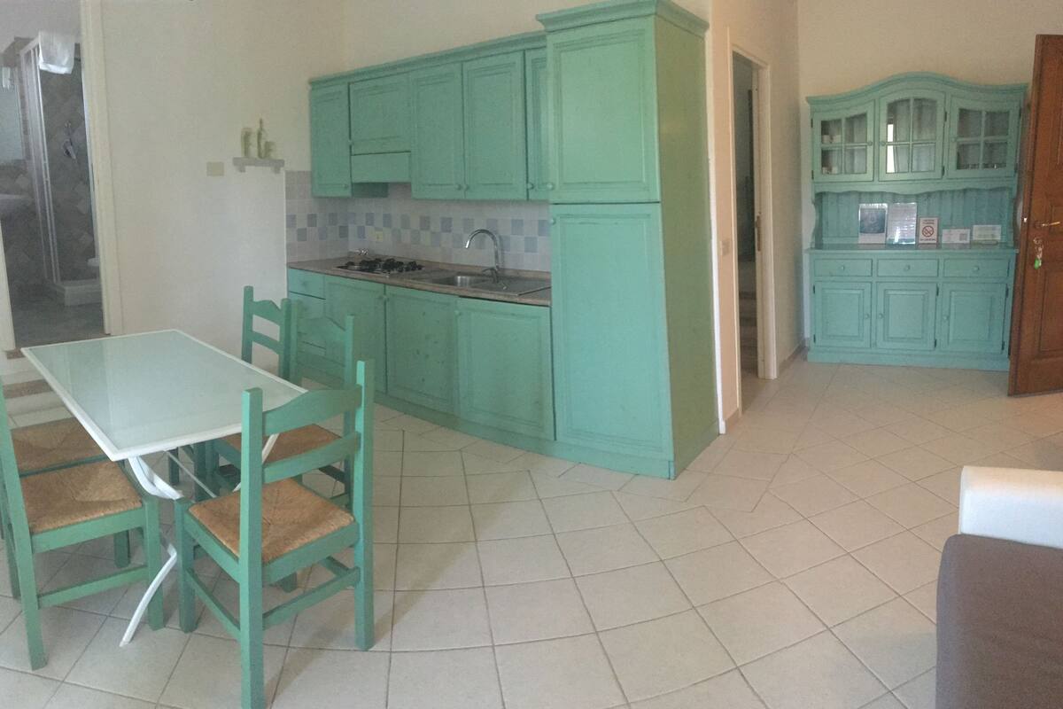 Une cuisine avec des armoires de couleur turquoise, un plan de travail blanc et une table en verre avec des chaises.