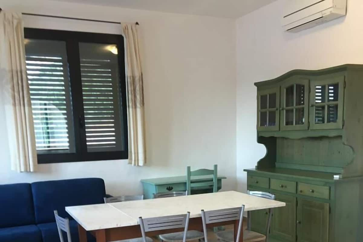 Un coin repas avec une table et des chaises, un canapé bleu, une fenêtre avec des stores et un meuble vert.
