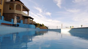 Una piscina con superficie d'acqua cristallina, una zona relax con sedie e un tavolo, e un edificio con facciata piastrellata e un balcone.