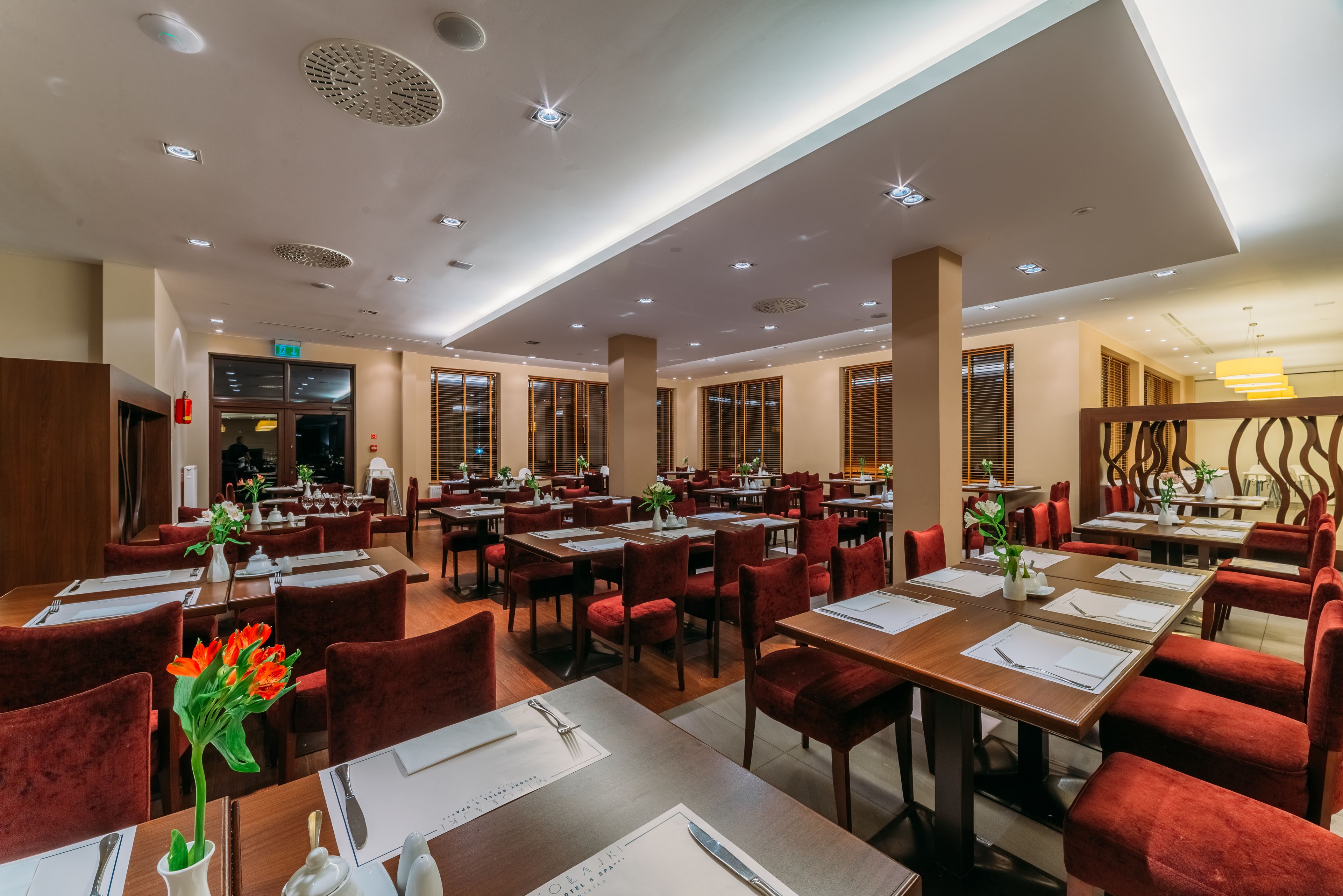 Een ruim restaurant met meerdere eettafels en rode bekleden stoelen, met een modern plafondontwerp en natuurlijk licht door grote ramen.