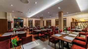 Een ruim restaurant met meerdere eettafels en rode bekleden stoelen, met een modern plafondontwerp en natuurlijk licht door grote ramen.