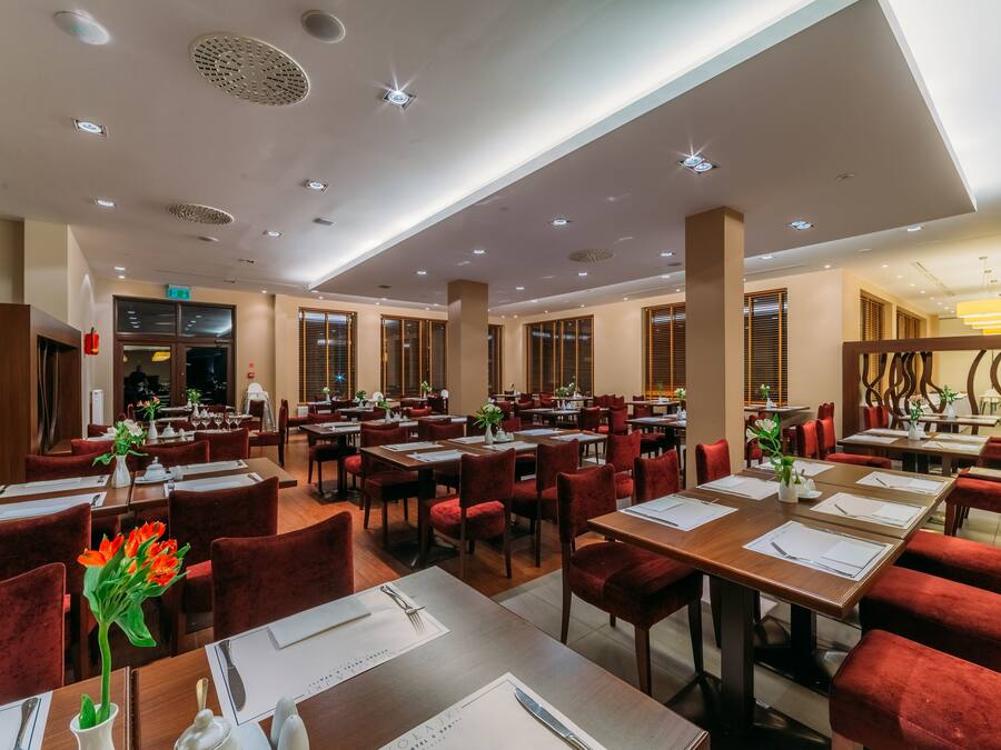 Een ruim restaurant met meerdere eettafels en rode bekleden stoelen, met een modern plafondontwerp en natuurlijk licht door grote ramen.