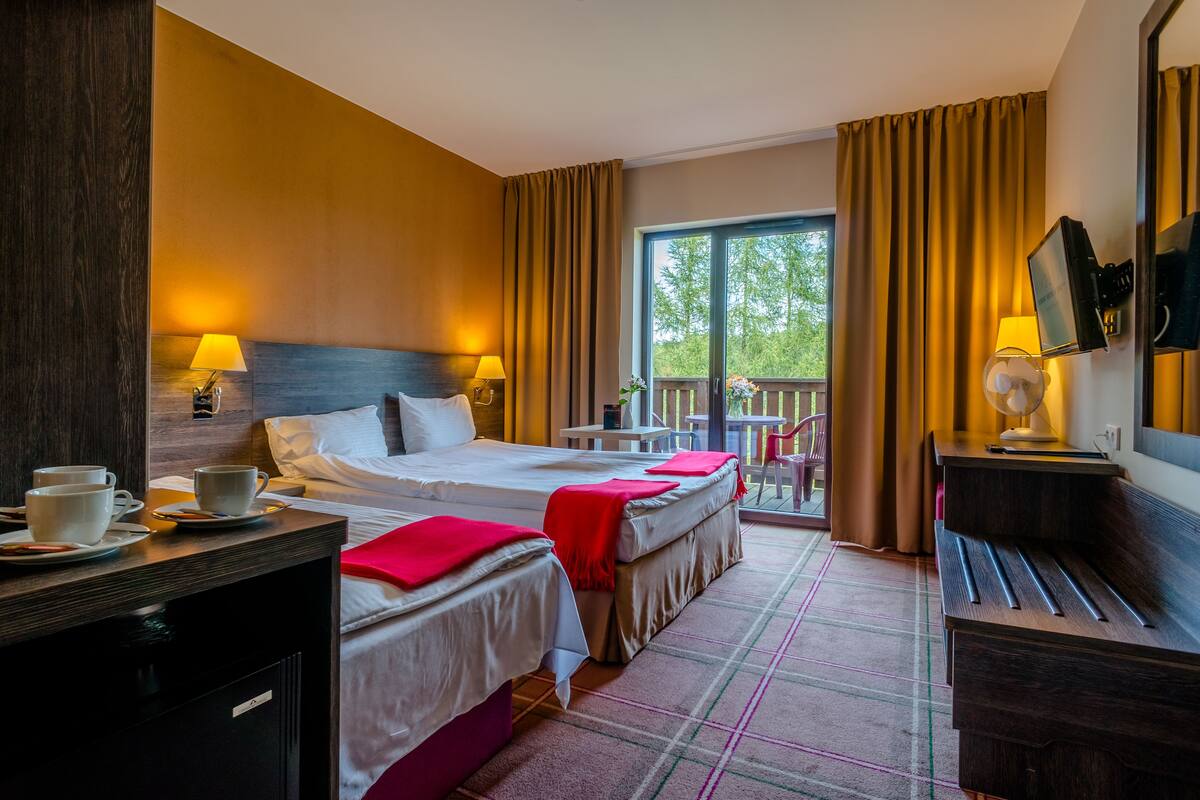 Quarto de hotel com duas camas, uma mesa com cadeira, uma mesinha e varanda com vista para árvores.