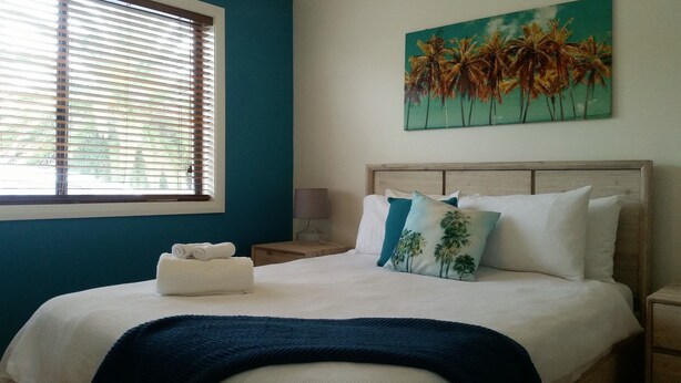 A bedroom with a bed, a window with blinds, a painting of palm trees, and a nightstand with a lamp. - Karuah Riverside Motel (Karuah)