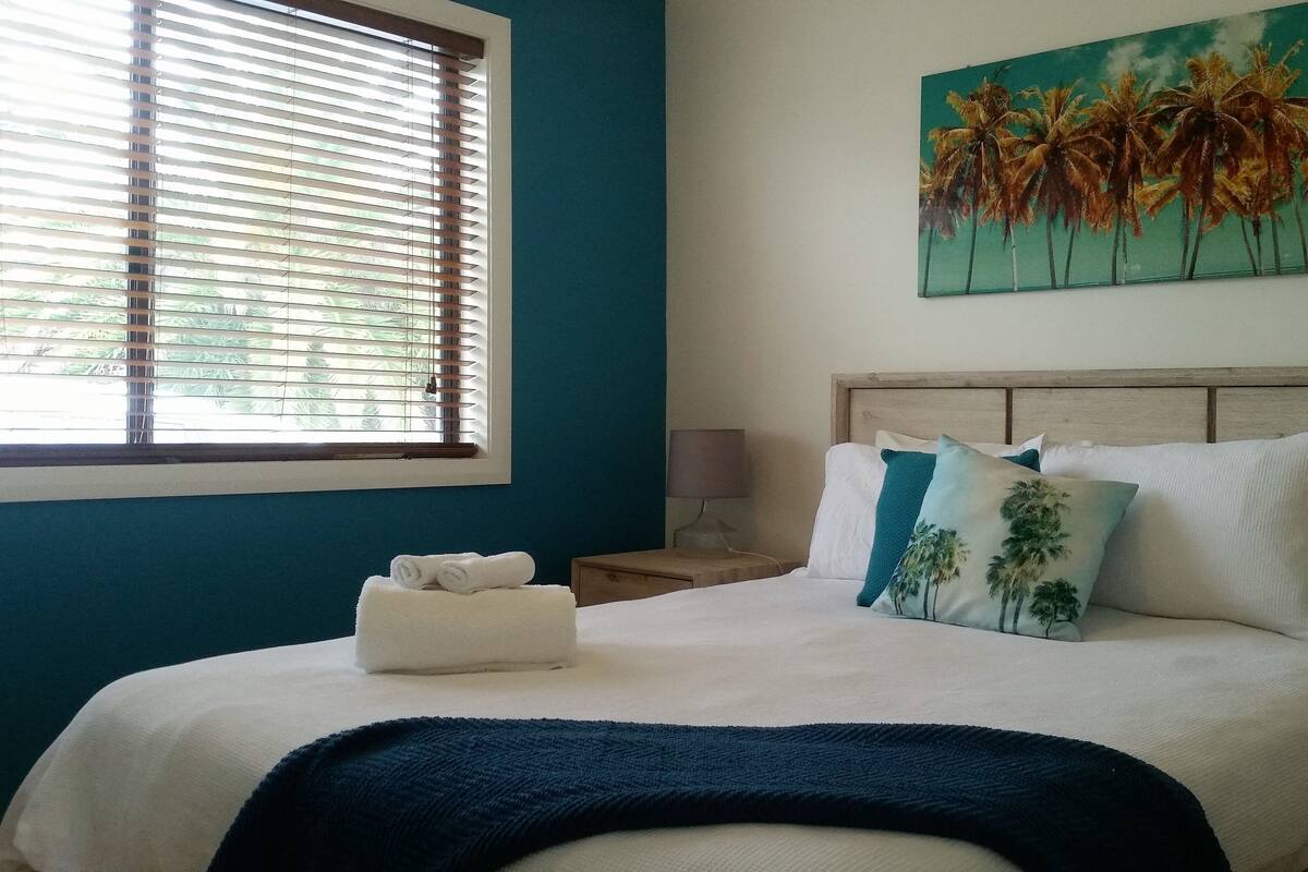 A bedroom with a bed, a window with blinds, a painting of palm trees, and a nightstand with a lamp.