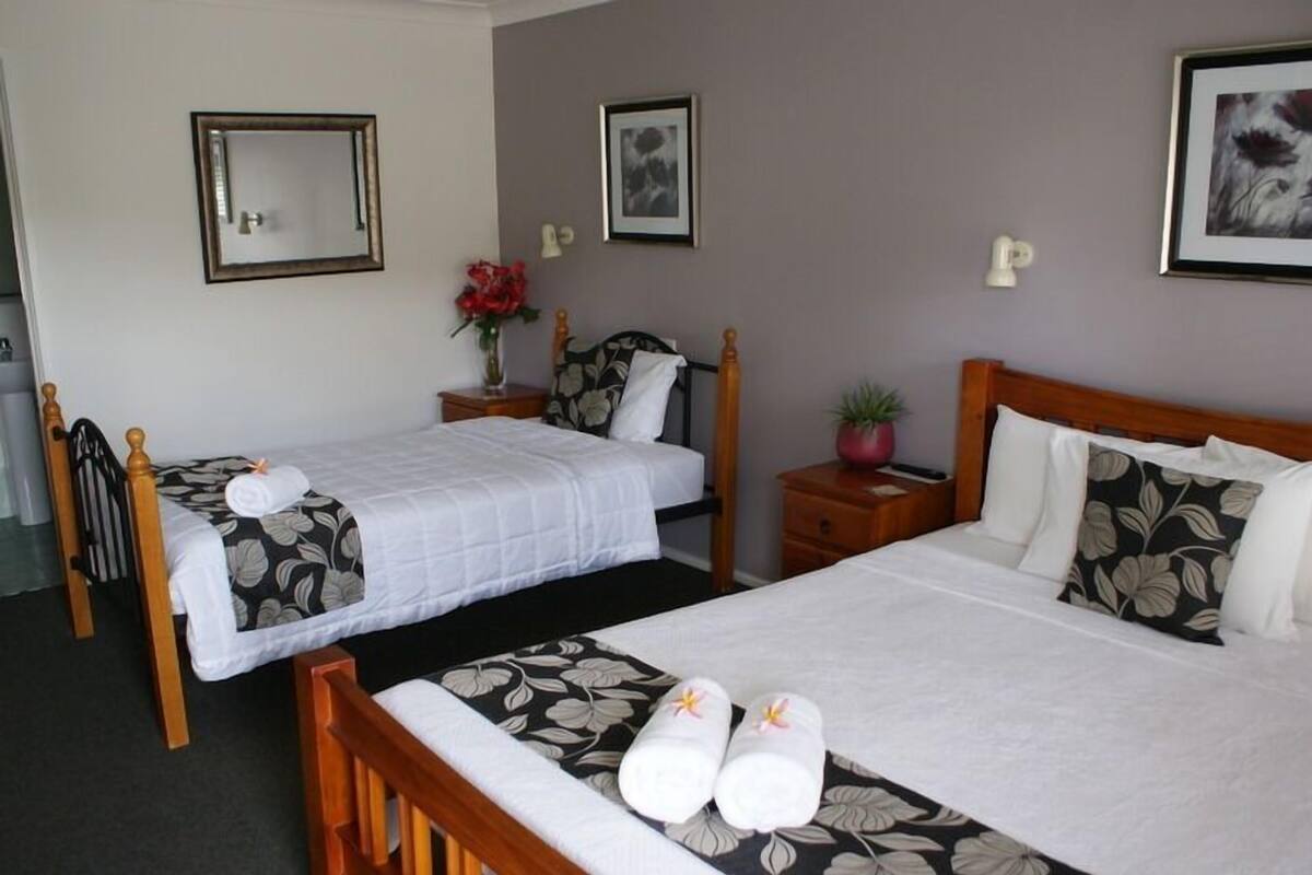 A hotel room with two single beds, a wooden headboard, a nightstand with a vase of flowers, and a framed picture on the wall.