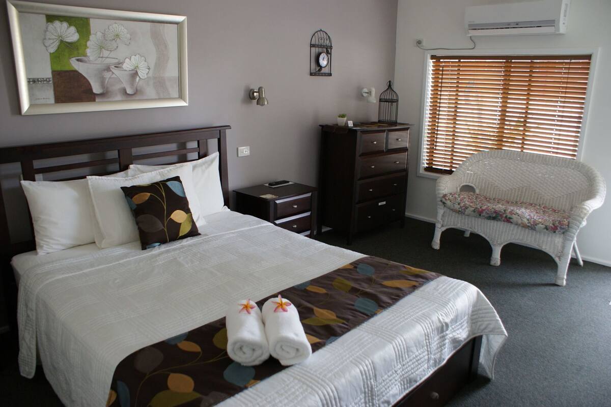 A neatly made bed with pillows, a bedside table, a dresser, a chair, and a wall-mounted clock.