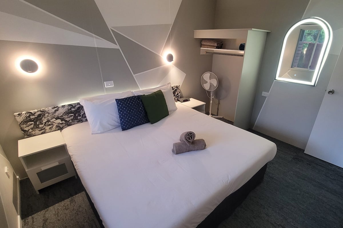 A modern hotel room with a large bed, a bedside table, a nightstand, a wall-mounted fan, a built-in shelf, and a small window with a light.