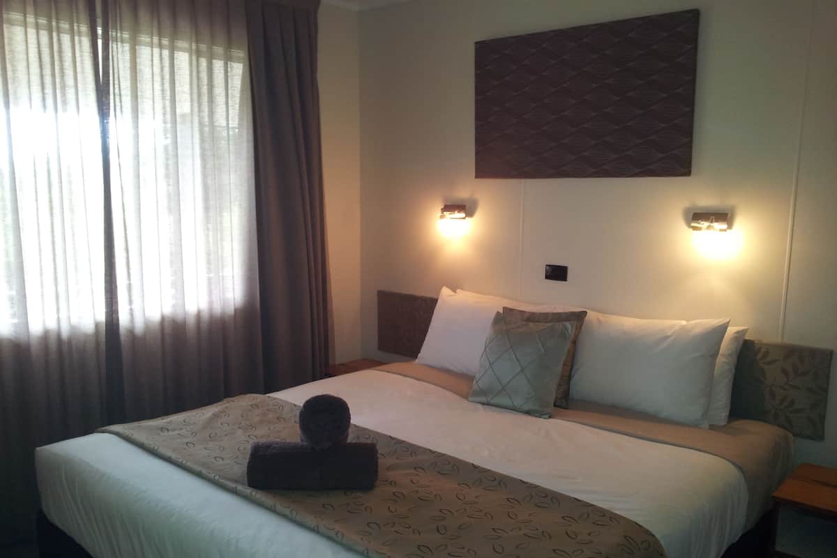 A hotel room with a large bed, two bedside lamps, a headboard with a leaf pattern, and a window with curtains.