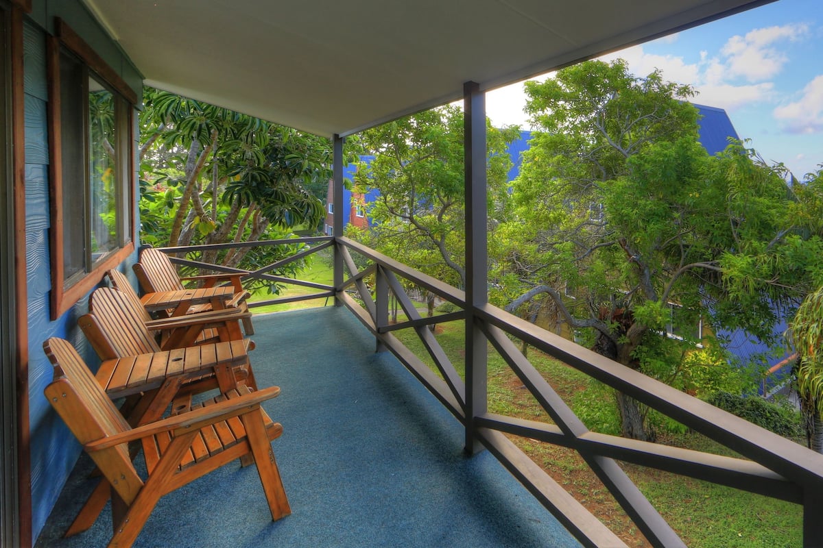A balcony with wooden chairs and a blue carpet, overlooking a lush garden and a building in the background.