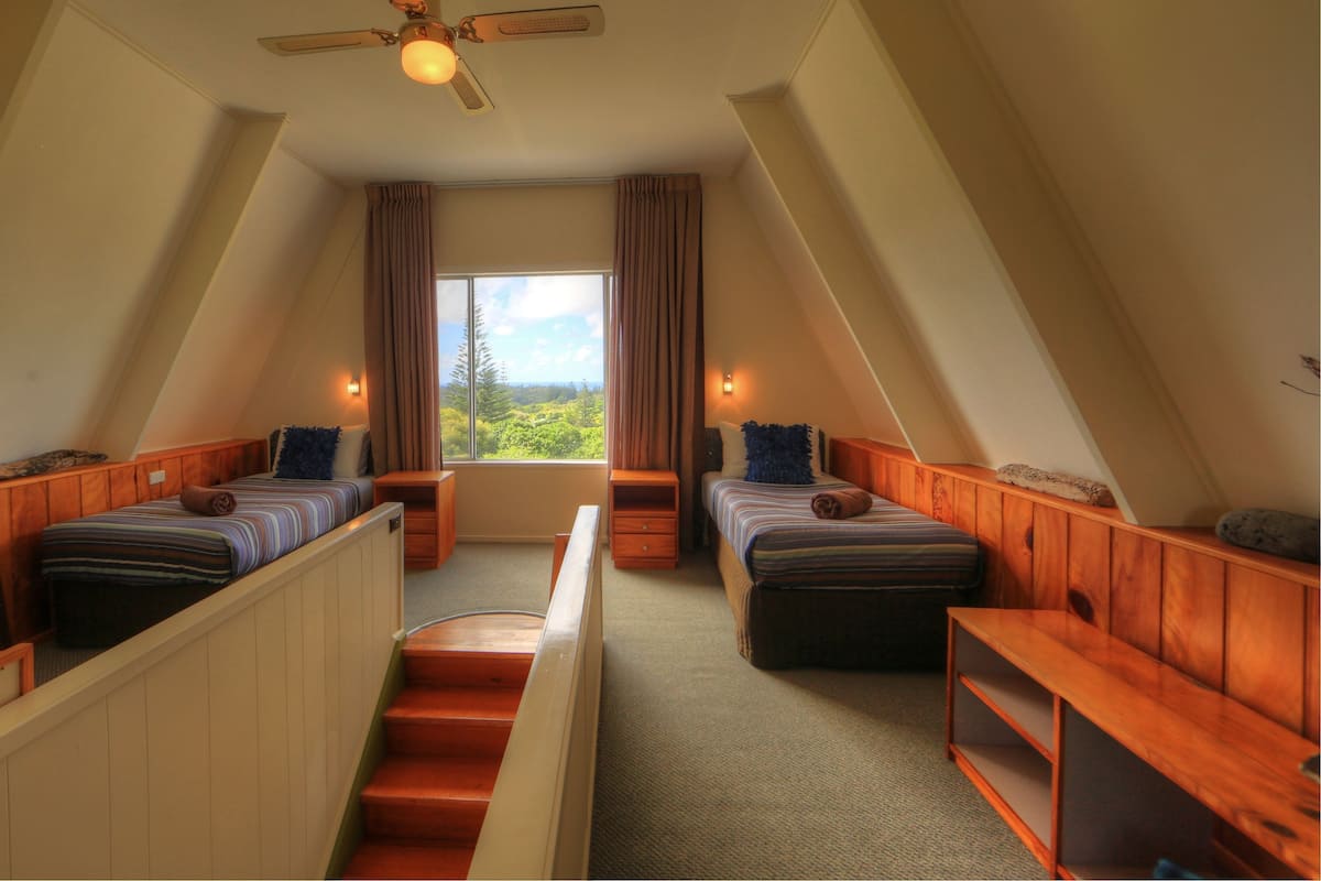 A cozy attic bedroom with two beds, a wooden staircase, and a large window offering a view of greenery.