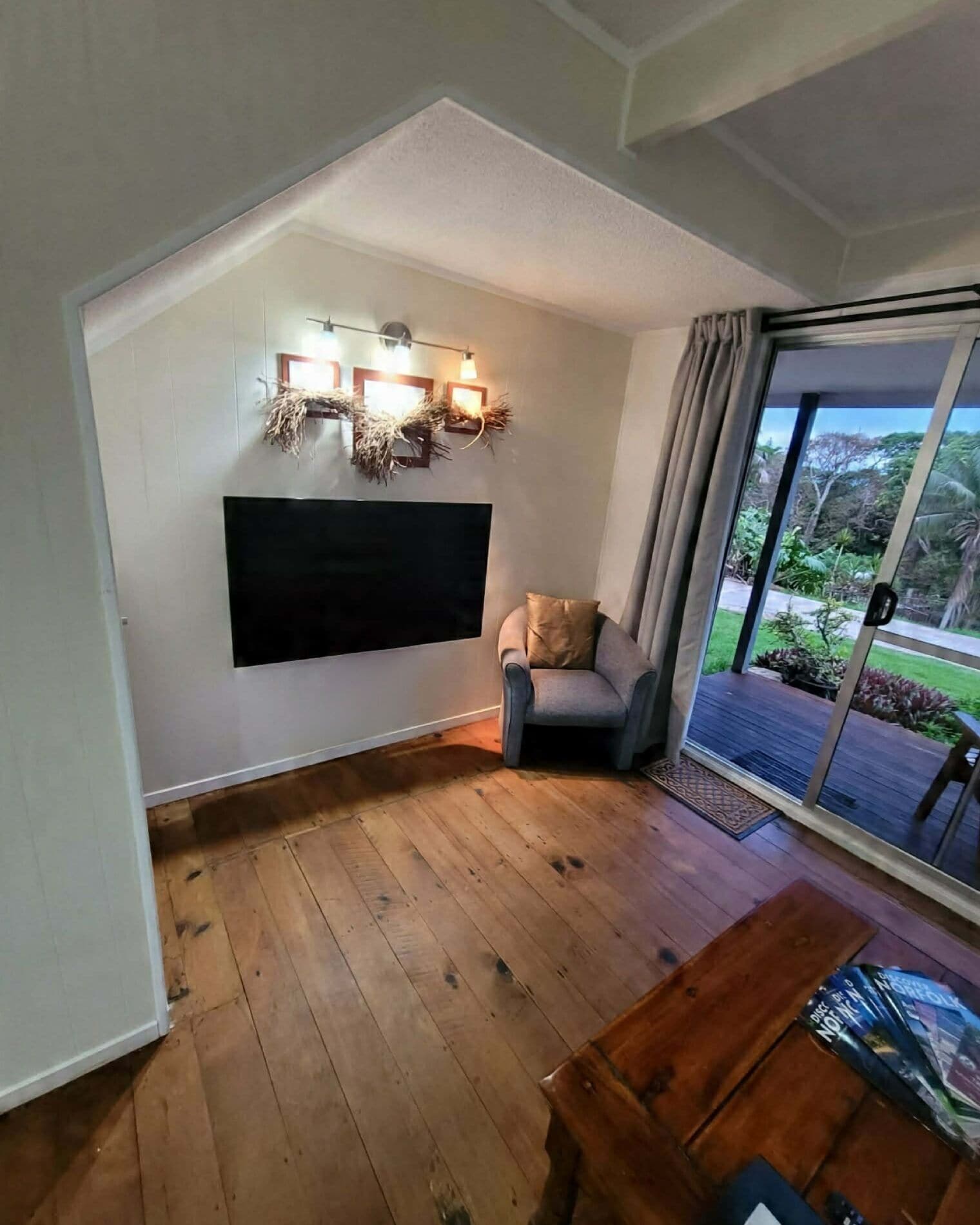 Una habitación con techo inclinado, un televisor de pantalla plana montado, un sillón gris, piso de madera y un amplio ventanal corredero que da a un espacio exterior con follaje.