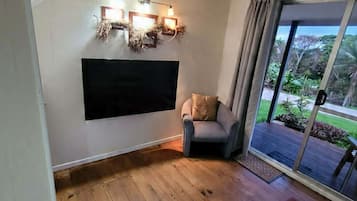 A room with a sloped ceiling, a mounted flat-screen TV, a grey armchair, a wooden floor, and a large sliding glass door leading to an outdoor area with greenery.
