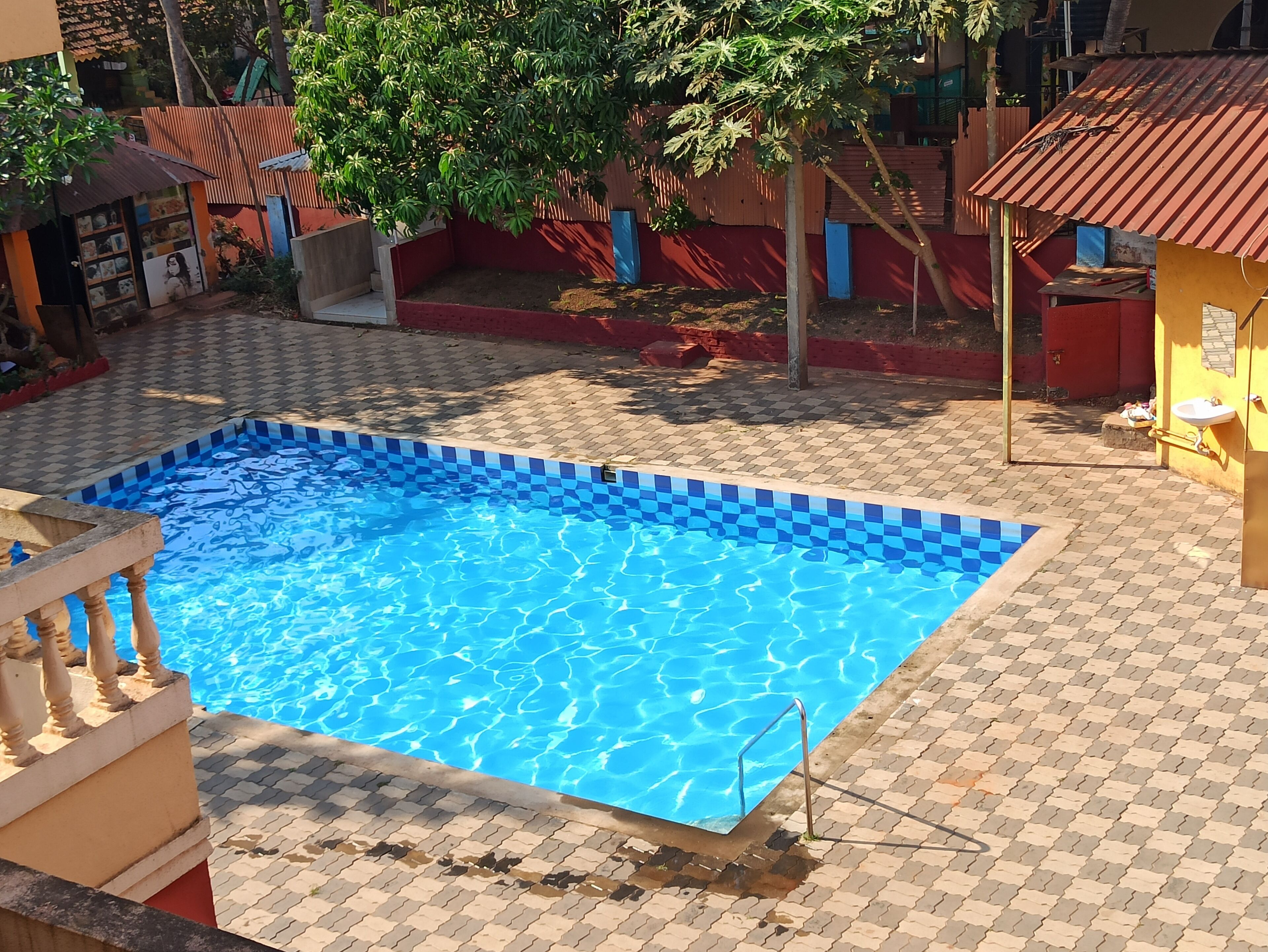 Ein Schwimmbecken mit Schachbrettmuster-Fliesenrand, umgeben von einer gepflasterten Fläche mit rotem und blauem Zaun.
