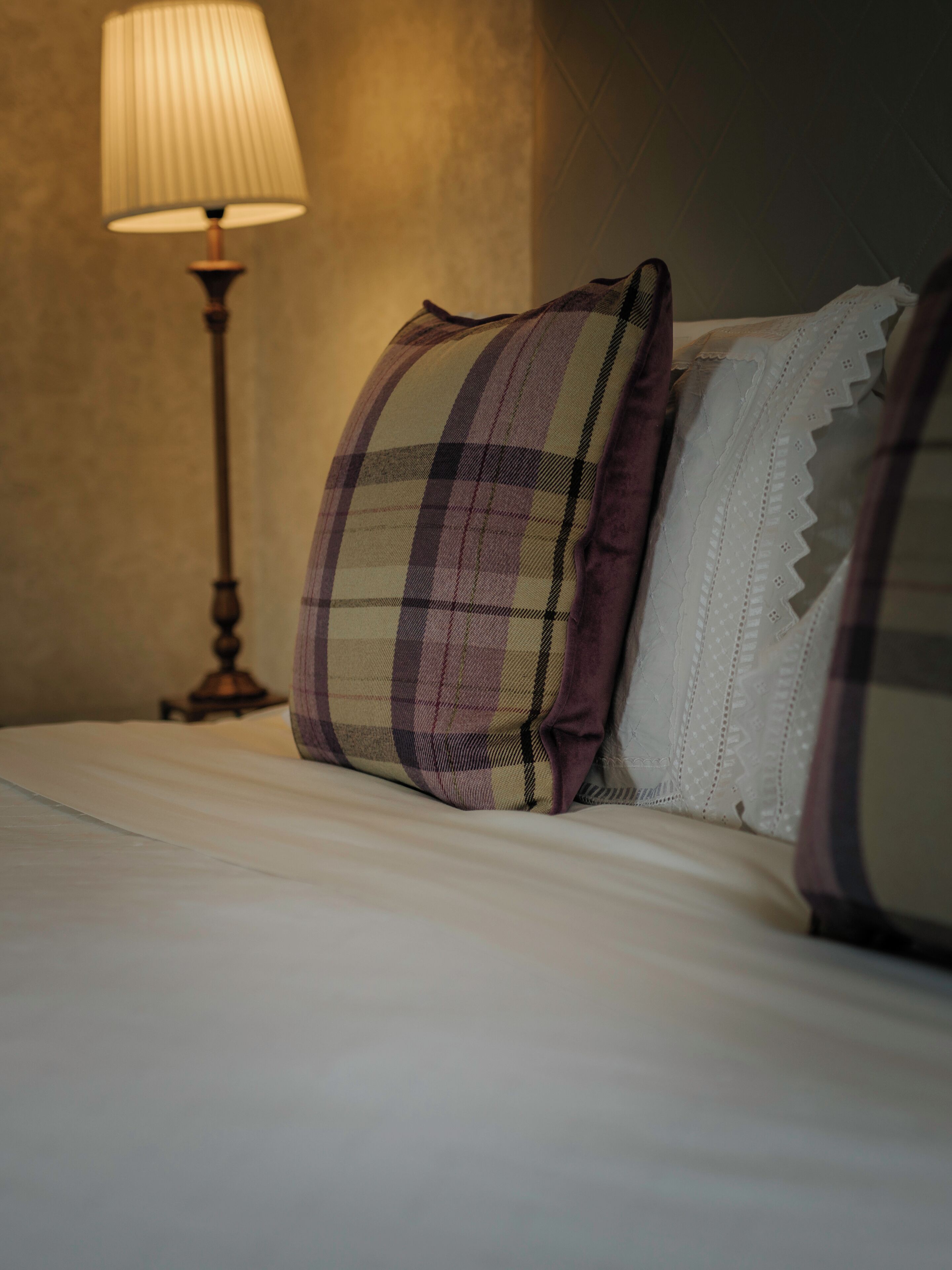 A neatly made bed with two pillows, one with a plaid pattern and the other with a lace trim. A table lamp is positioned to the left of the bed.
