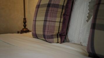 A neatly made bed with two pillows, one with a plaid pattern and the other with a lace trim. A table lamp is positioned to the left of the bed.