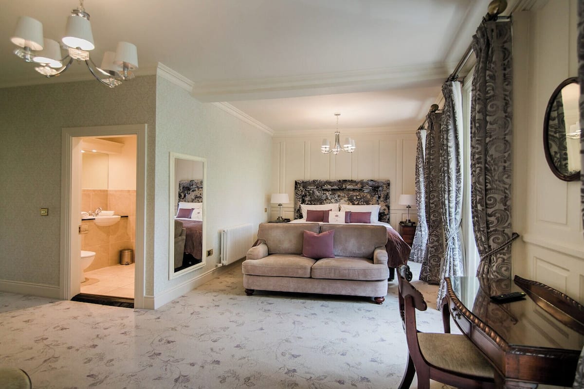 A hotel room with a large bed, a sitting area with a sofa, a mirror, and a chandelier.