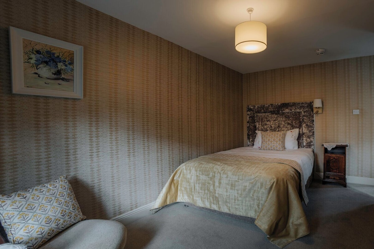 A bedroom with a bed, a nightstand, a lamp, a chair, and a painting on the wall.