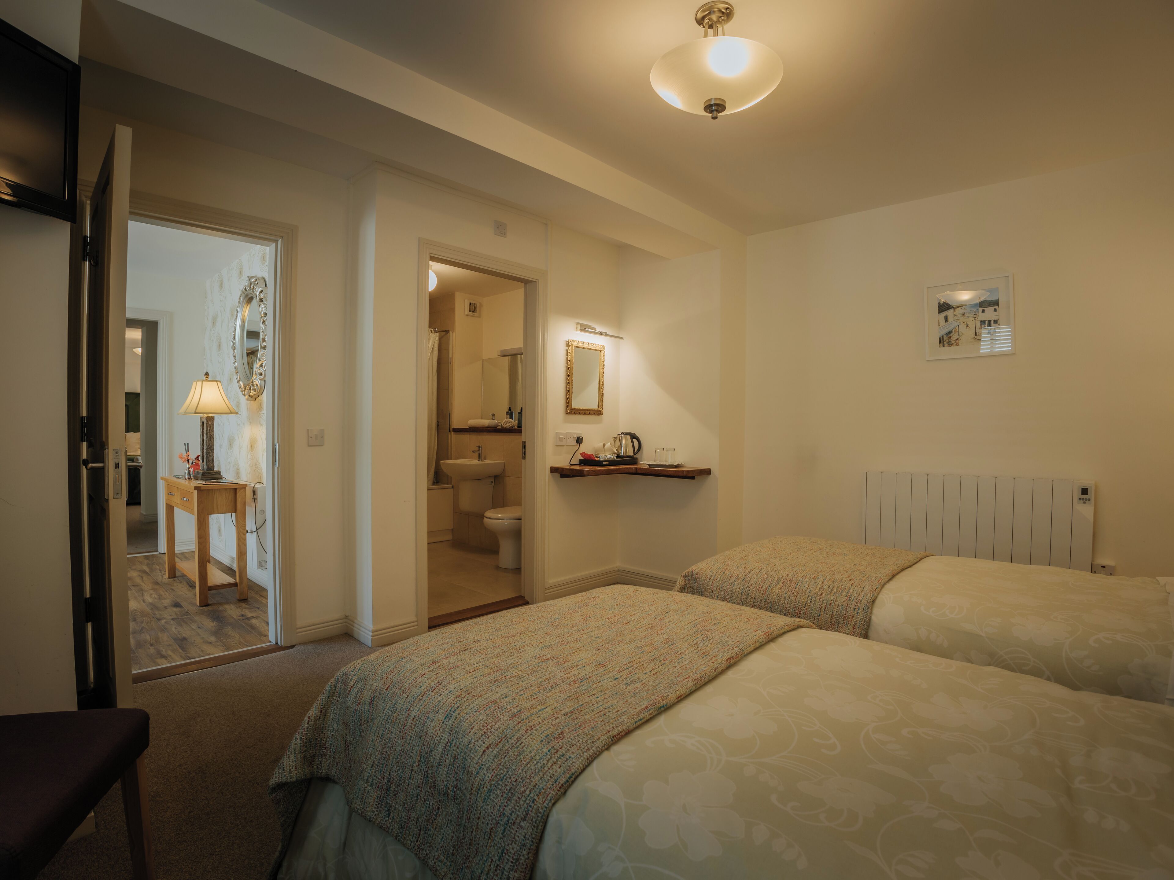 A hotel room with two beds, a bedside table with a lamp, a bathroom visible through an open door, and a radiator on the wall.