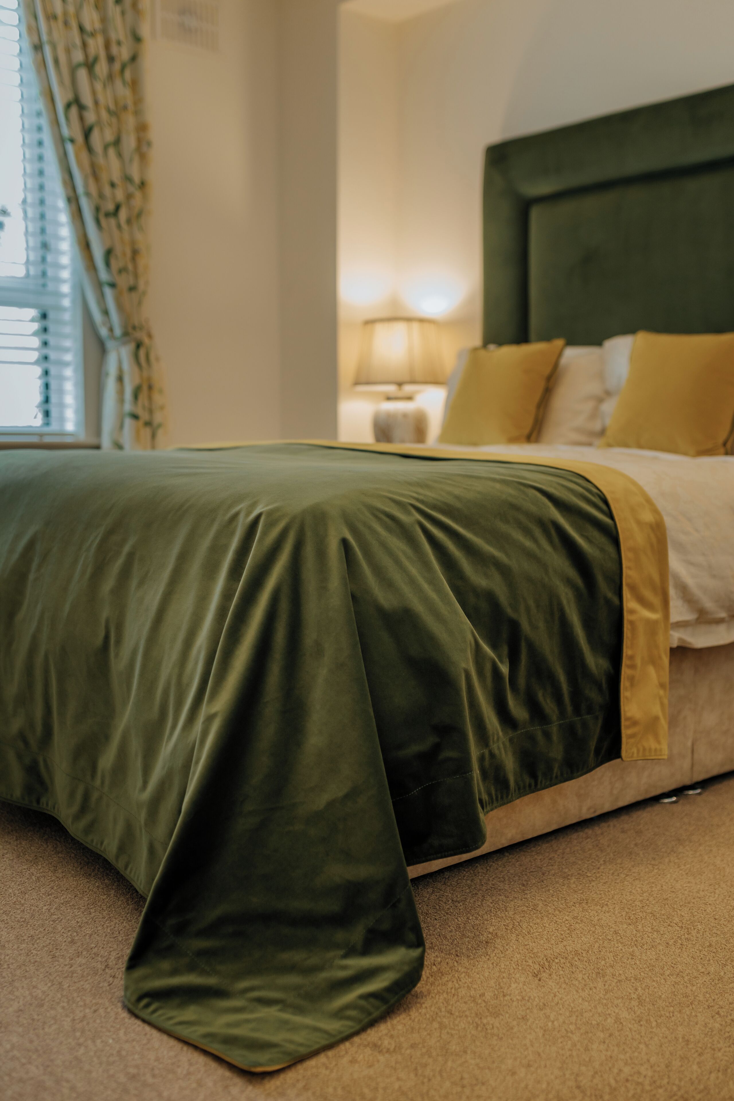 Una cama bien tendida con una colcha verde y almohadas amarillas, una lámpara de noche y una ventana con cortinas.