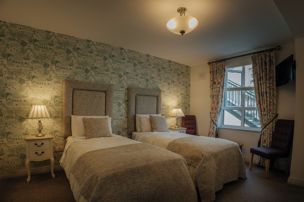 A bedroom with two beds, a nightstand, a chair, a window with curtains, and a television on the wall.