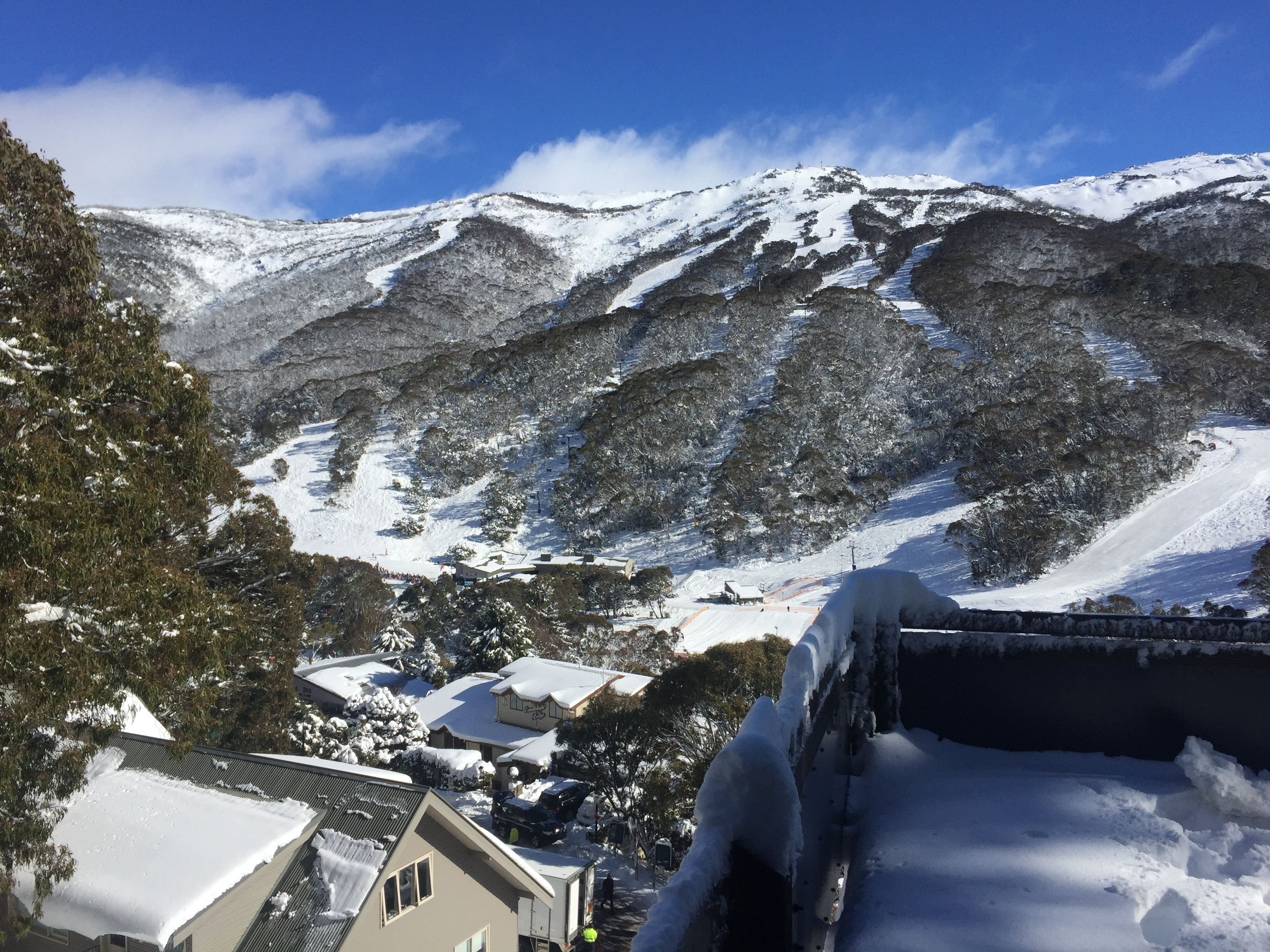 Paisagem montanhosa coberta de neve, com área residencial em primeiro plano, casas com neve nos telhados e céu azul límpido.