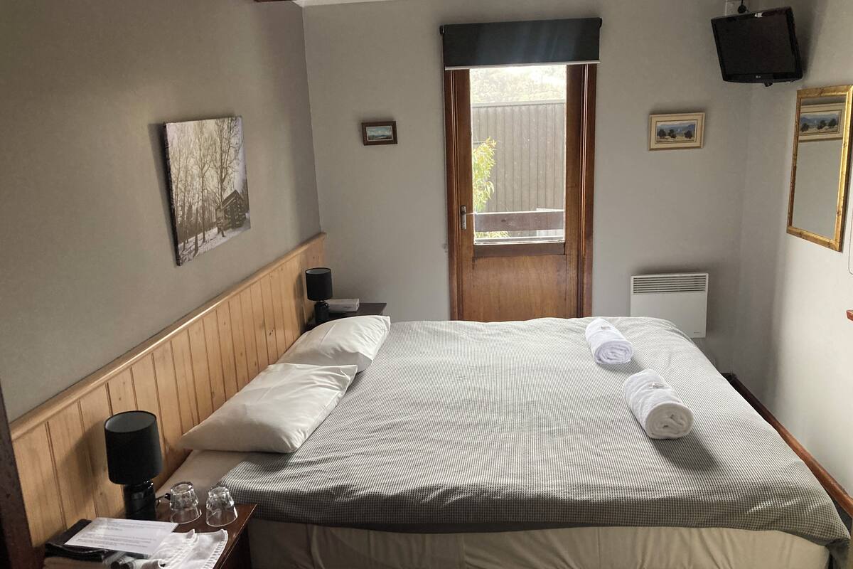 Ein ordentlich bezogenes Bett mit einer gesteppten Decke, eine Nachttischlampe, ein kleiner Tisch mit einem Glas, ein Handtuch und ein gerahmtes Bild an der Wand.