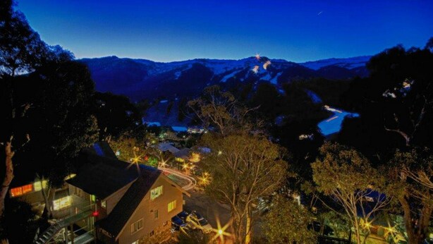Paisaje montañoso nocturno con una casa en primer plano y un río al fondo.