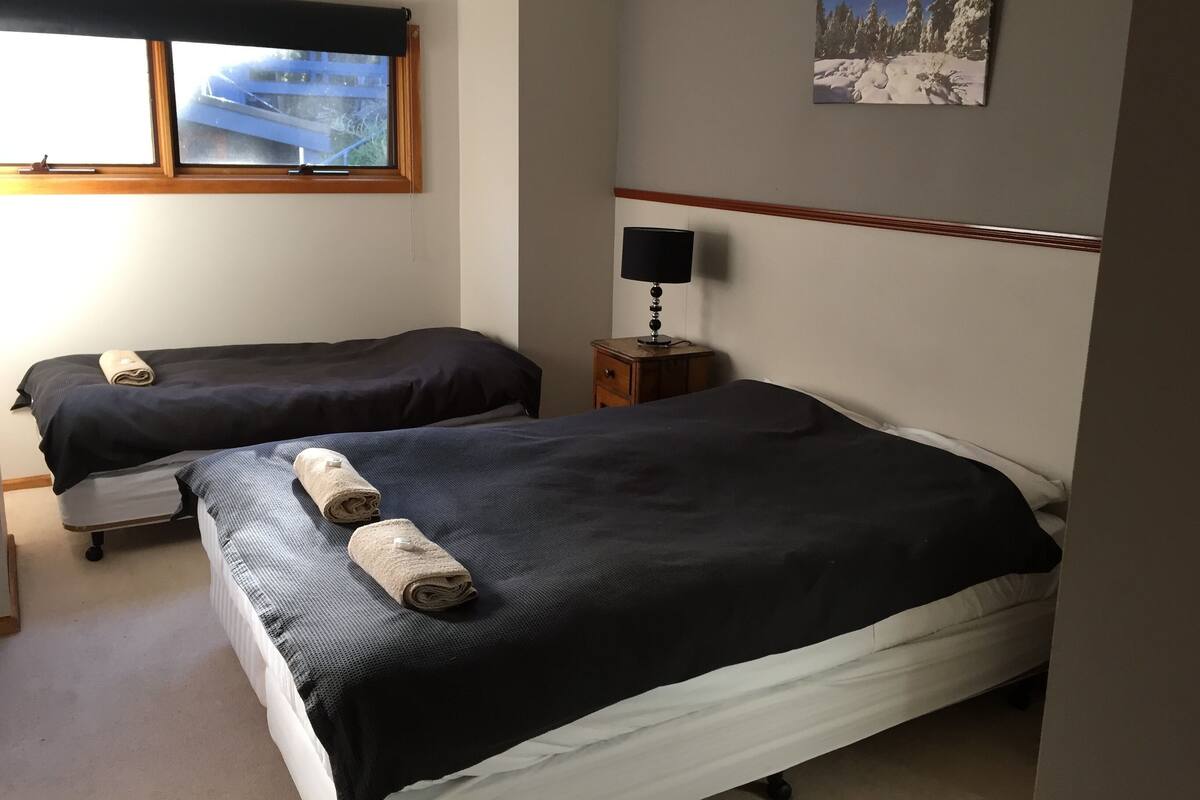 Ein Hotelzimmer mit zwei Betten, einem Nachttisch mit Lampe, einem Fenster mit Blick auf ein blaues Gebäude und einem Bild einer verschneiten Landschaft an der Wand.