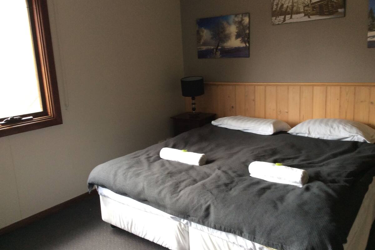Ein ordentlich bezogenes Bett mit grauer Decke und weißen Kissen, ein Holz-Kopfbrett, ein Nachttisch mit Lampe und ein Fenster mit Jalousien.