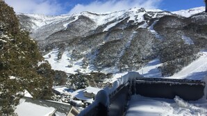 Un paisaje nevado de montaña con una estación de esquí en el valle, que incluye edificios y un telesilla.
