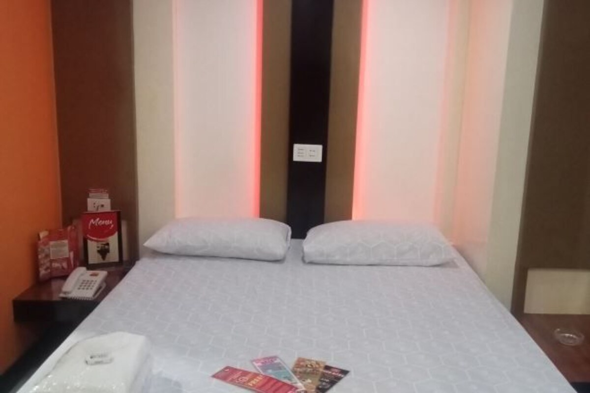 Une chambre d’hôtel comprenant un lit, des oreillers, une table de chevet avec un téléphone et une boîte, et un mur orné de bandes lumineuses verticales.