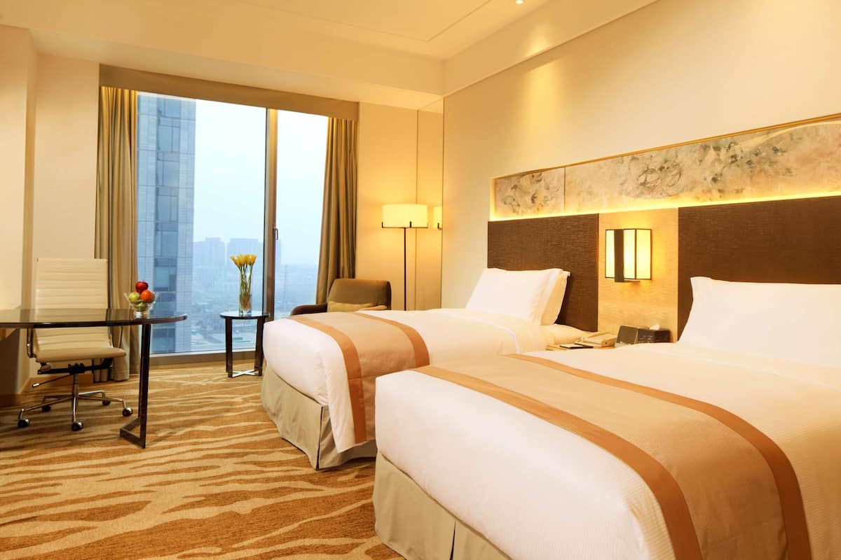 Une chambre d’hôtel avec deux lits, un bureau et une chaise. De grandes fenêtres offrent une vue sur le paysage urbain.