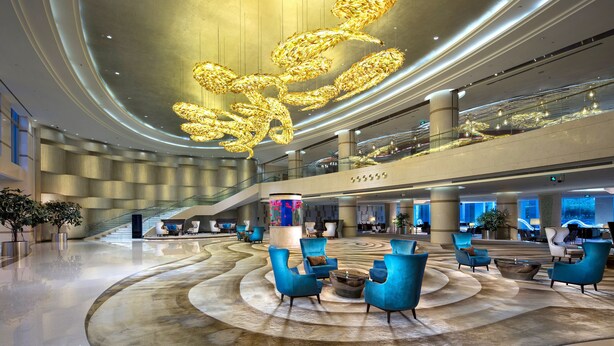 Um hall amplo com design moderno, apresentando uma área de estar central com cadeiras azuis, uma grande escultura decorativa no teto e uma escadaria que dá acesso aos andares superiores.