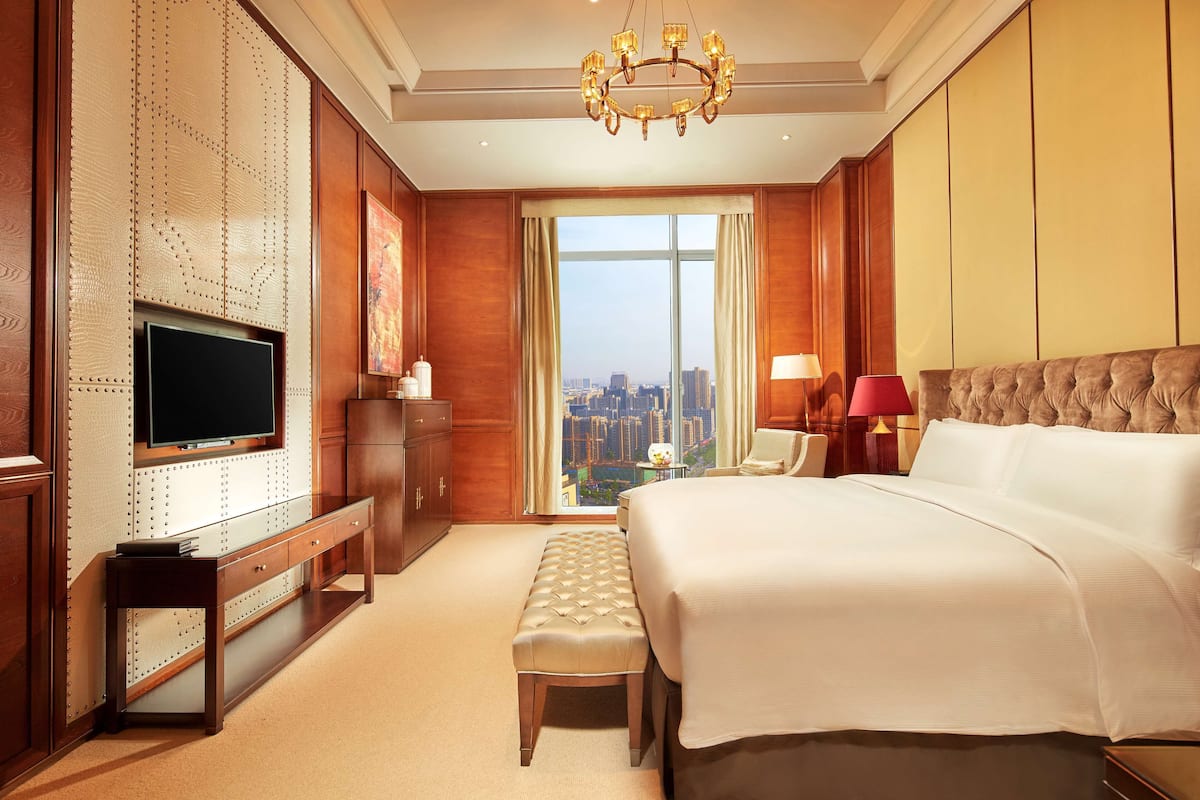 Une chambre d’hôtel avec un grand lit, une télévision, un banc et une vue sur le paysage urbain, visible à travers une grande fenêtre.