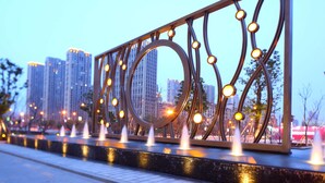 Uma moderna fonte ao ar livre com estrutura metálica geométrica e iluminação, com o horizonte da cidade ao entardecer.