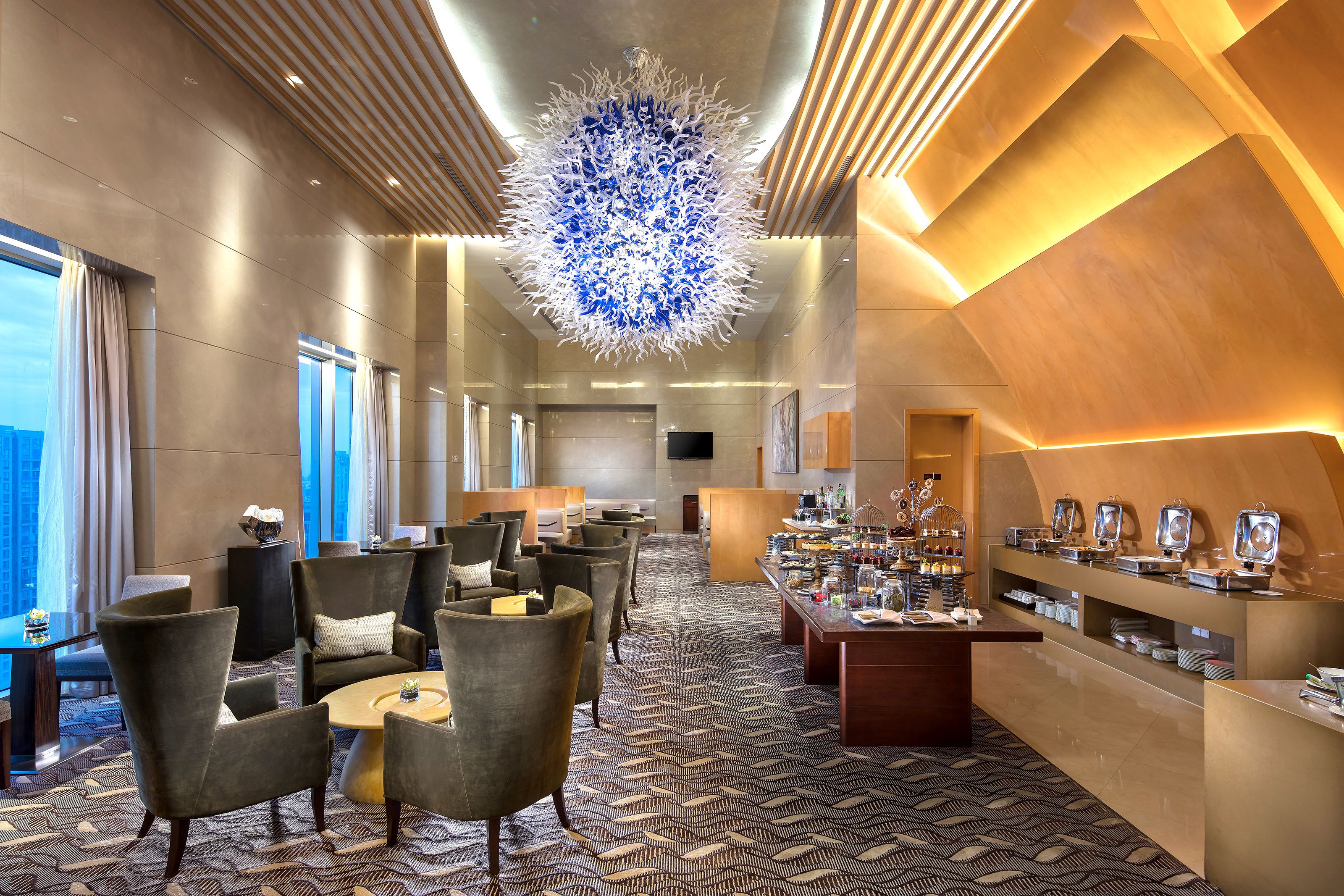 A modern hotel lobby with a central chandelier, seating area, and buffet.