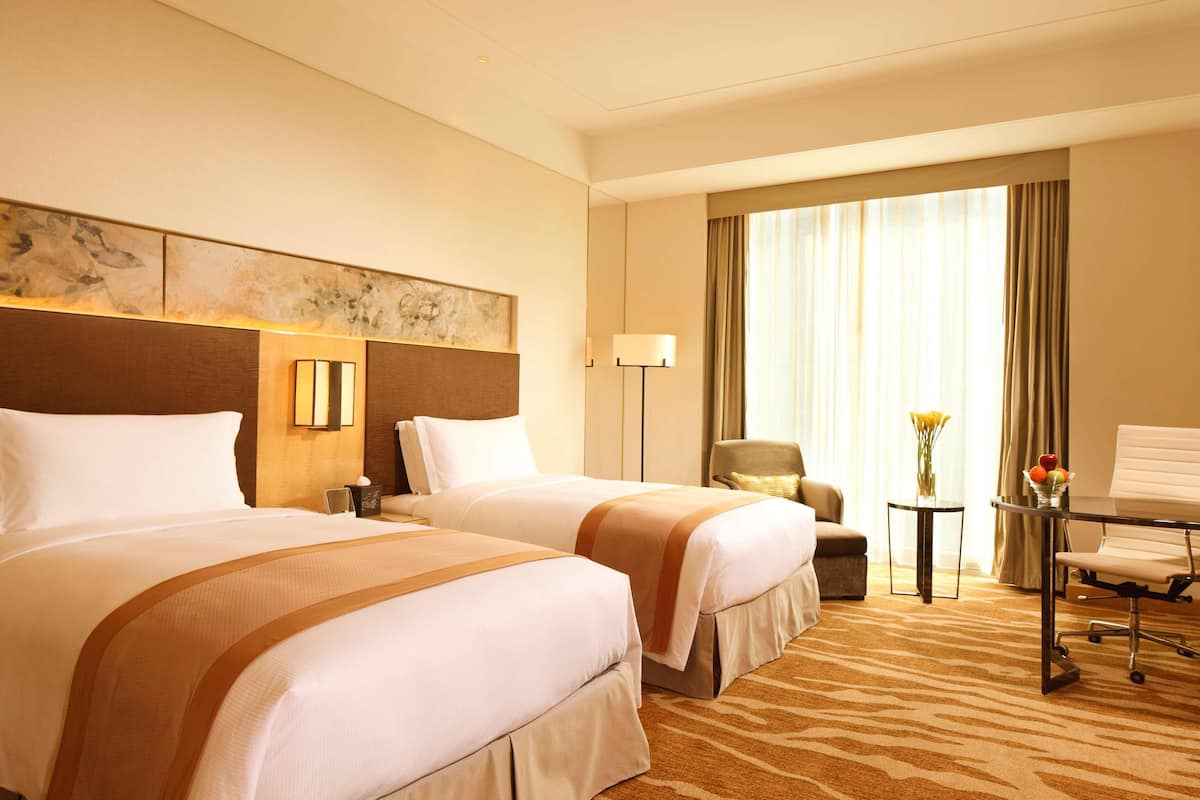 Une chambre d’hôtel avec deux lits, un bureau, une chaise, une lampe et une fenêtre avec des rideaux.
