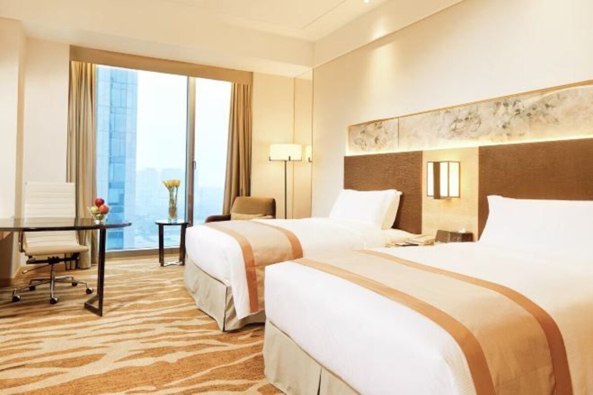 A hotel room with two beds, a desk, a chair, a lamp, and a view of a cityscape.