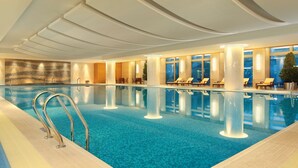Uma grande piscina interior com água azul cristalina, rodeada por espreguiçadeiras e um design de teto moderno.