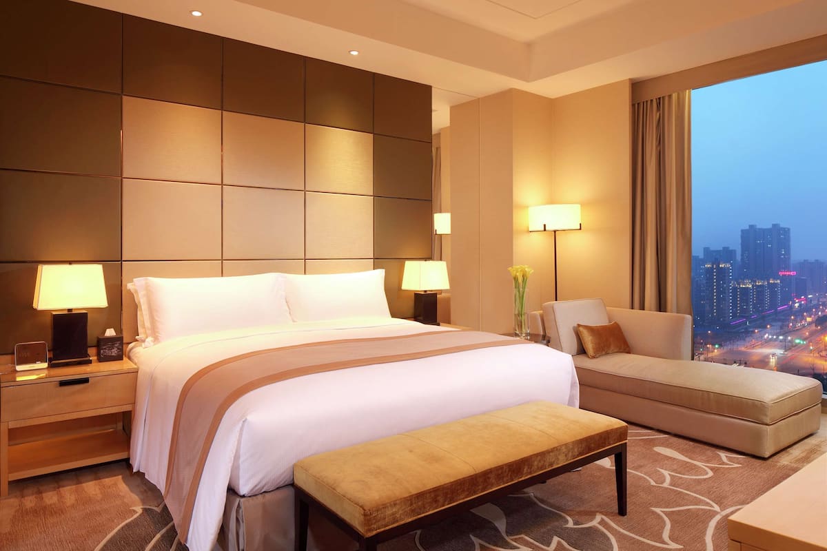 Une chambre d’hôtel avec un grand lit, des tables de chevet avec des lampes, un canapé et une vue sur la ville la nuit.