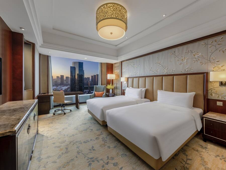 A hotel room with two beds, a desk, and a chair. There is a large window with a cityscape view.