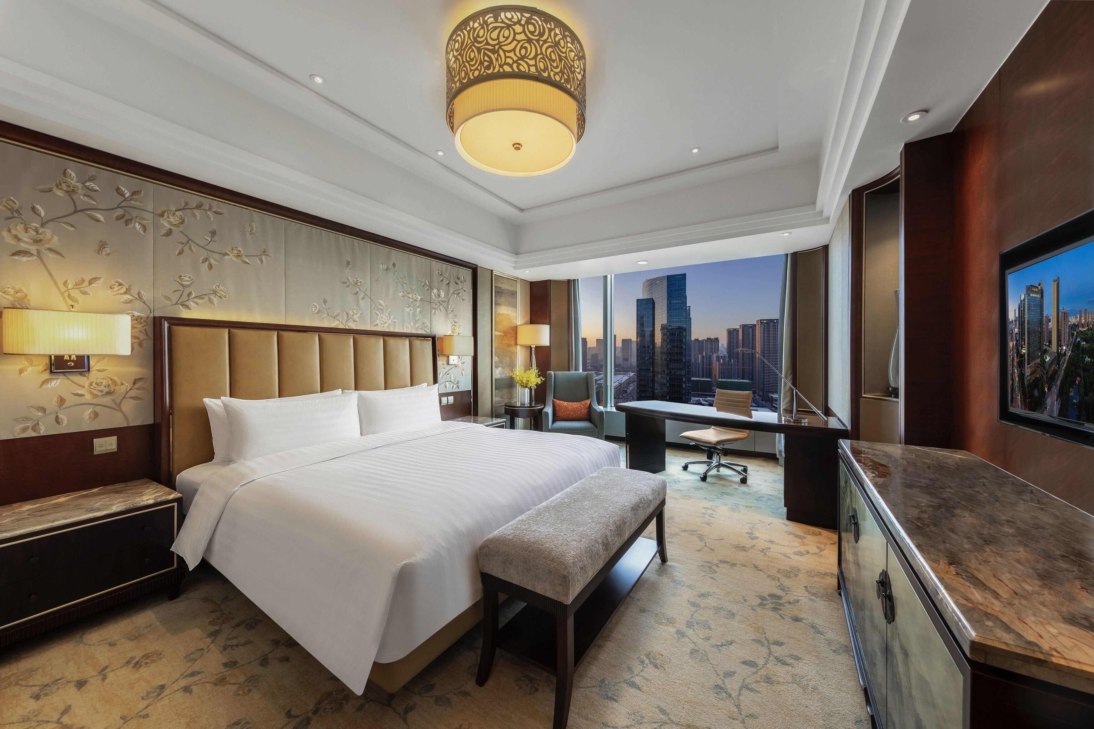 Quarto de hotel com cama grande, escrivaninha, cadeira, banqueta, televisão e vista para a cidade.